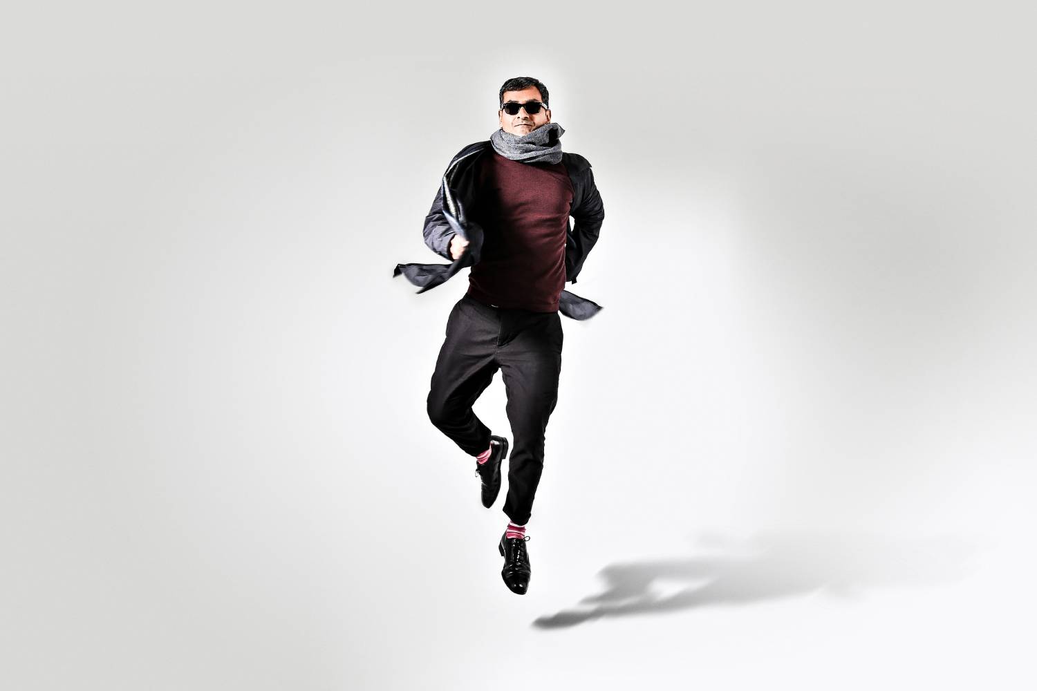 An image of Harry jumping into the air in front of a grey background. He is wearing black trousers, a red jumper and a grey scarf with black sunglasses..