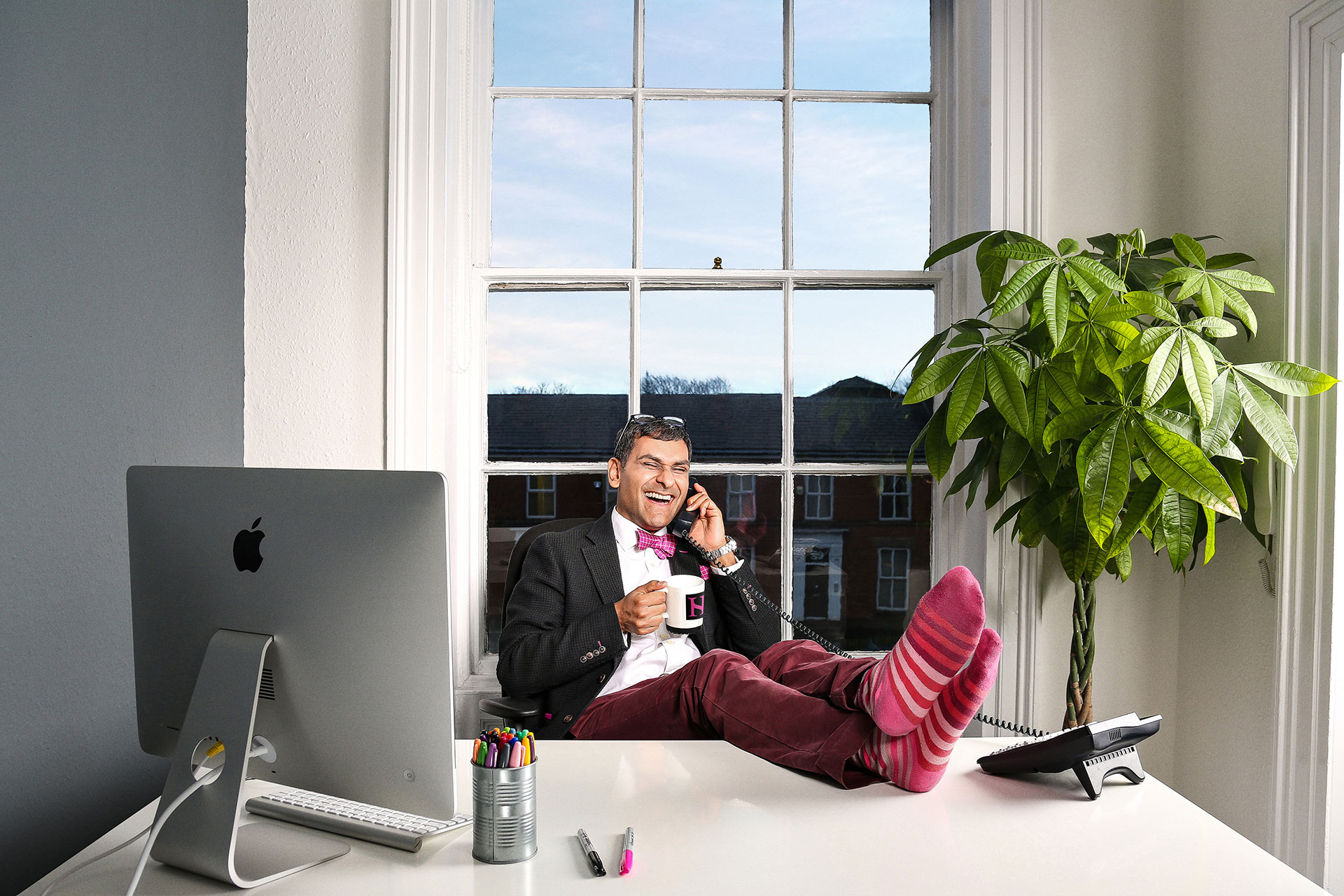 An image of Harry sat behind a white desk, leaning back with his feet propped up on the desk. He is laughing on the phone, holding a mug.