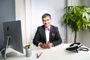BF1A0775 An image of Harry Suleman wearing a red and pink patterned shirt. He is wearing a red bowtie and is fixing it whilst smiling at the camera.